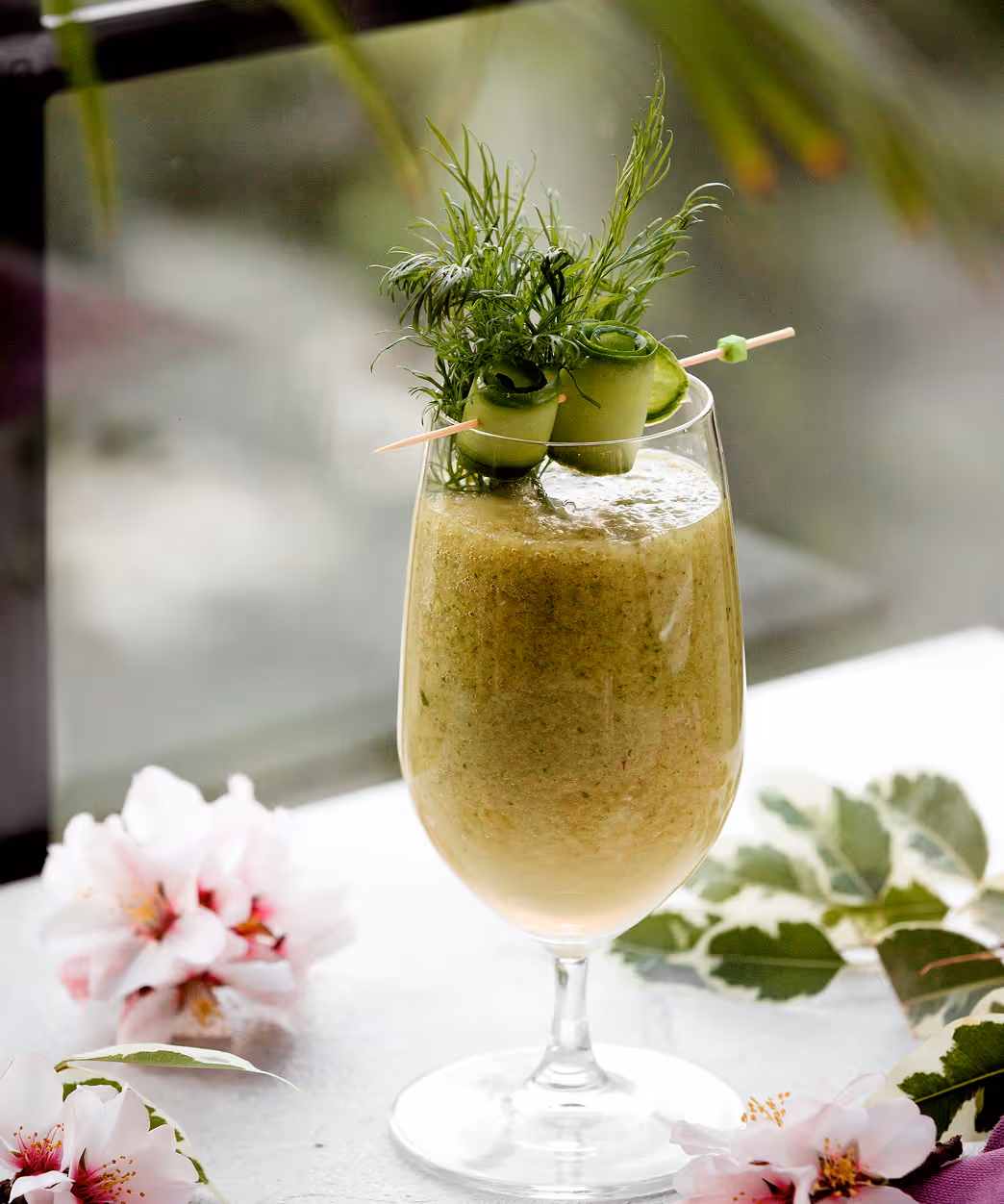 Tall glass of green smoothie garnished with dill and cucumber rolls on a skewer, surrounded by pink flowers and green leaves.